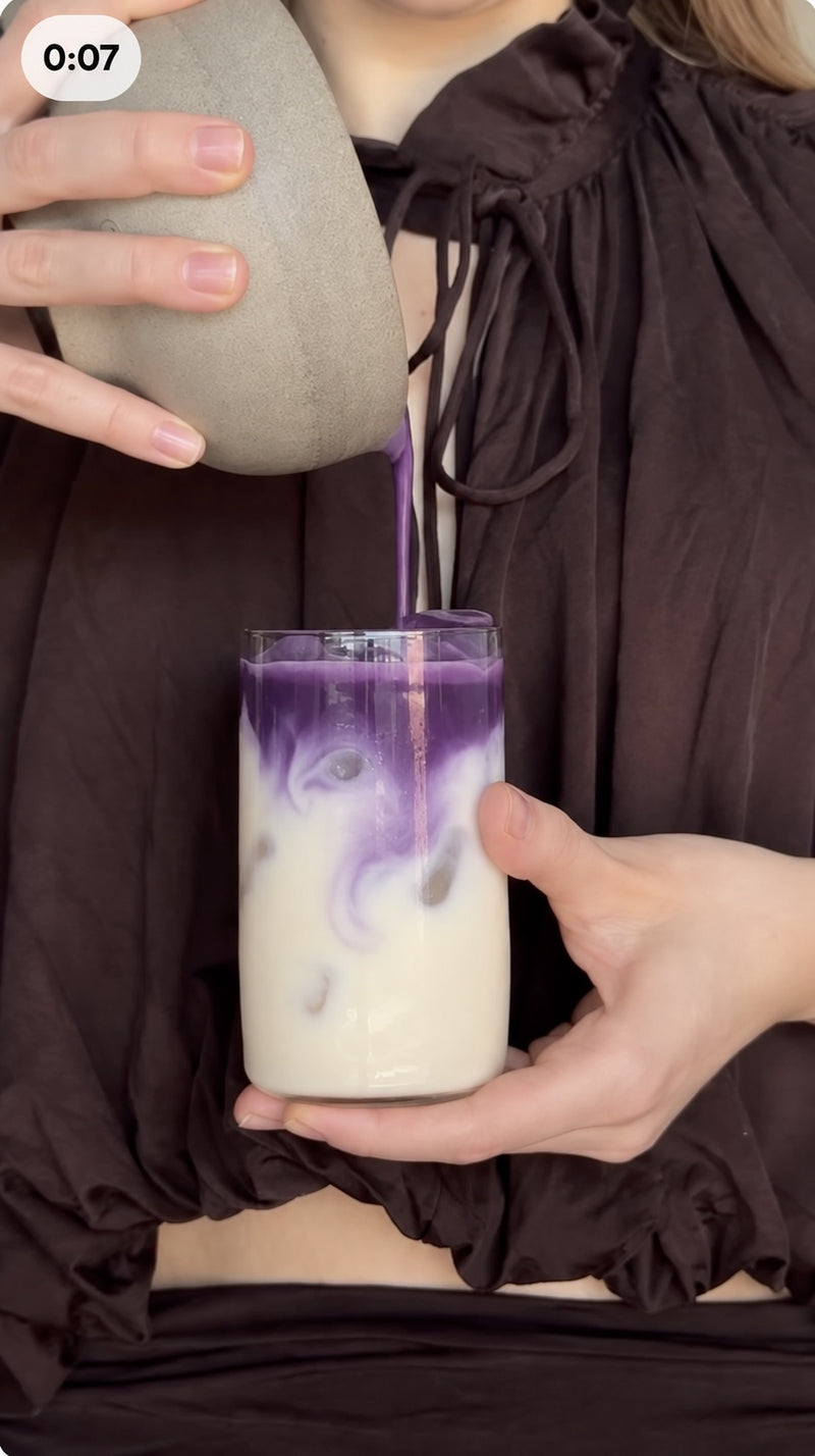 Femme préparant un ube latte glacé en versant une boisson violette dans un verre de lait, rituel Vytal Oya sans caféine.